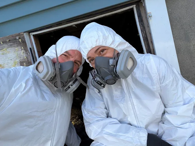 Two technicians in white hazmat suits and respirators entering crawl space for pest control treatment