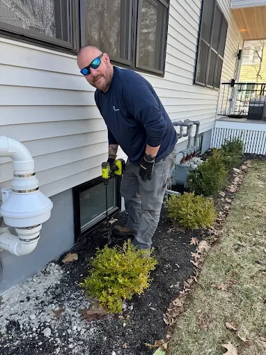Pest control technician treating foundation perimeter with spray equipment near vinyl siding and landscaping